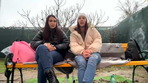 Media: Video of two women sitting on a park bench, dressed in winter coats, under bare trees, with a pink backpack and green fence in the background.