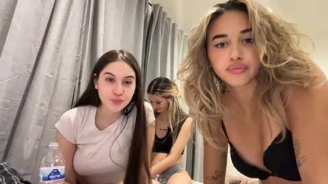 Media: Video of three young women with light skin tones, long hair, and casual clothing. They are in a bedroom with gray curtains, one with a bottle, another with a tattoo.