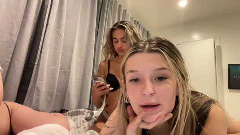 Media: Video of a young blonde woman in a black bra, lying on a bed, looking at her phone, while another blonde woman, also in a bra, rests her chin on her hand.