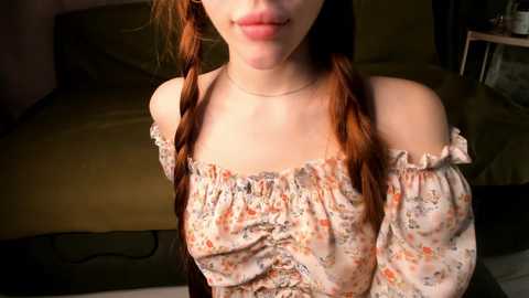 Video of a young woman with pale skin and long, reddish-brown hair in pigtails, wearing a floral off-the-shoulder blouse, sitting indoors on a green couch with a wooden table and chair in the background.