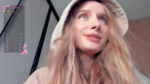 Media: Video of a young Caucasian woman with long, wavy blonde hair, fair skin, and green eyes, wearing a beige bucket hat and light-colored top. Background includes a black metal frame and a pink, floral digital filter.