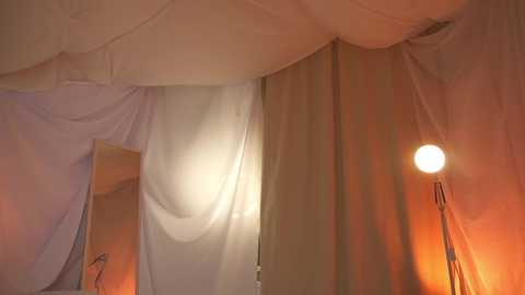 Media: Video of a dimly lit room with sheer white curtains draped over a bed, illuminated by a single warm-toned lamp on the right, creating a cozy, intimate atmosphere.