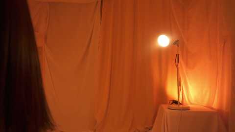 Media: A video of a dimly lit, cozy room with warm orange and yellow hues. A round table with a white cloth holds a vintage lamp casting a soft glow. The background features sheer, draped curtains.