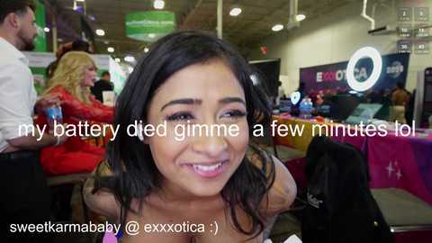 Media: Video of a smiling woman with dark hair, wearing a pink top, at a busy electronics trade show, with a text overlay reading \"my battery died gimme a few minutes lol.\