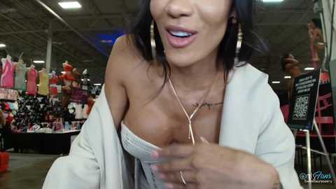 Media: A video shows an Asian woman with dark hair, wearing a white cardigan over a low-cut white dress, and a gold necklace. She's smiling in a bustling indoor market with colorful clothes and signs in the background.