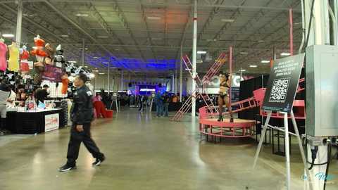 Media: Video of a brightly lit, industrial warehouse with a man walking left, a woman on a pink stage right, and clothing racks and banners in the background.