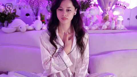 Media: Video of an Asian woman with long black hair, wearing a white silk robe, sitting on a white couch. Background features a soft purple ambiance with decorative flowers and plush toys.