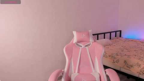 Video of a pink gaming chair with white accents, positioned in a simple, beige-walled bedroom with a floral-patterned bedspread and a black metal headboard.