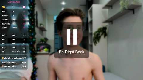 Media: Video of a shirtless young man with short dark hair, blurred background, \"Be Right Back\" text overlay, social media app interface, and Christmas decorations.