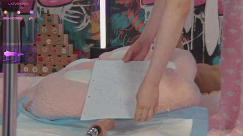 Media: A video of a person with fair skin and long, straight, pink hair lying on a fluffy pink bedspread, using a white clipboard. The background features colorful, abstract artwork and a wooden box.