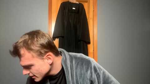 Media: Video of a man in a gray robe, leaning forward, with a black robe hanging in the background against a gray wall.