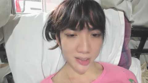 Media: Video of a young Asian woman with short dark hair, lying on a hospital bed with white sheets, wearing a pink shirt. She appears calm and focused.