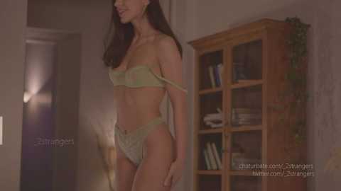 Media: A video of a slender, light-skinned woman with long dark hair, wearing a delicate, pale green lace bra and panties, standing in a dimly lit room with a wooden bookshelf and plants in the background.