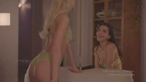 Media: A video depicts a blonde woman in a green lace lingerie set leaning over a table, smiling at a brunette woman with a similar outfit in a warmly lit room.