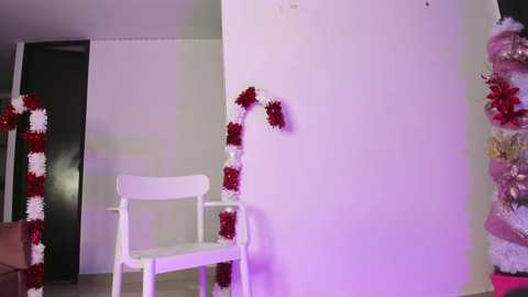 Media: A video of a minimalist room with a white chair, a red and white tinsel garland, and a purple light. The walls are white, and a black door is visible in the background.