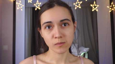 Media: Video of a young woman with fair skin and dark hair, wearing a light pink tank top, against a softly lit room with star-shaped lights and grey curtains.