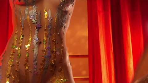 Media: Video of a partially nude woman with a medium brown complexion, adorned with a glittering body paint design of gold and purple floral patterns. She stands against a backdrop of vibrant red curtains, with a warm, orange glow in the background.