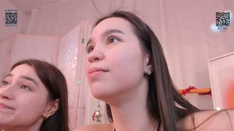 Media: Video of a young woman with long dark hair, light skin, and small earrings, looking contemplative in a softly lit room with a mirror, door, and wall-mounted QR code.