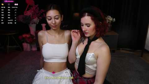 Media: Video of two young women, one with dark hair and a white crop top, the other with red hair and a black tie, in a dimly lit room with flowers.