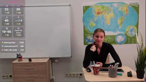 Media: Video of a young woman with light skin and brown hair in a black top, seated at a desk in a classroom. Background includes a large world map, a whiteboard, and a wooden cabinet.