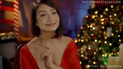 Media: Video of a smiling Asian woman with short black hair, wearing a low-cut red dress, standing in front of a Christmas tree with warm lighting.