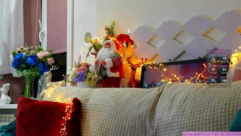 Media: A festive video of a cozy living room featuring a beige couch with a red throw, adorned with a large Santa figurine, Christmas lights, a bouquet of flowers, and a digital clock.