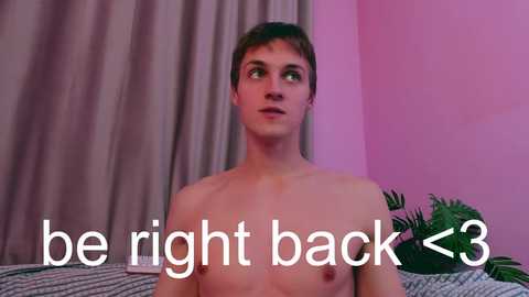 Media: Video of a shirtless young man with light skin and short brown hair, looking up. Text overlay: \"be right back <3\". Pink walls, green curtains, and a plant in the background.