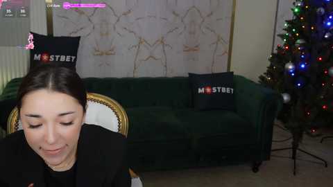 Media: Video of a woman with dark hair, wearing a black top, sitting on a green velvet sofa with two pillows reading \"MOSTBET\" and \"MOSTBET.\" A decorated Christmas tree stands in the background.