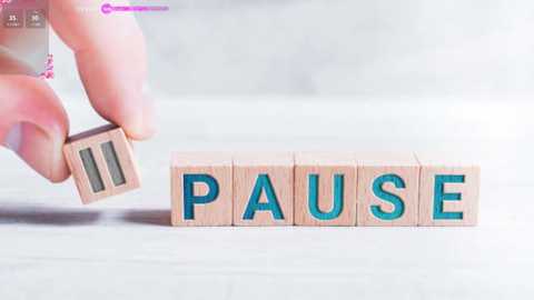 Media: Video of a hand holding a wooden block with \"PAUSE\" in blue letters, placed on a light wooden floor. The background is blurred with soft pastel hues.