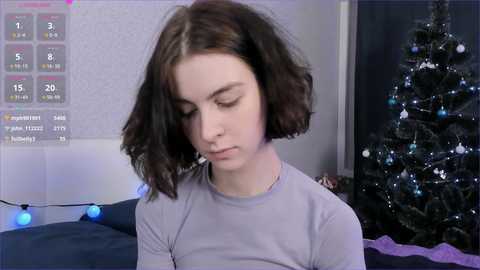 A young woman with shoulder-length brown hair sits in a dimly lit room, wearing a light gray shirt. In the background, a decorated Christmas tree with twinkling lights and ornaments stands beside a bed.