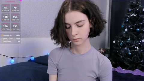 Media: Video of a young woman with shoulder-length, wavy dark hair, wearing a light gray t-shirt, standing in a cozy room with a decorated Christmas tree and blue string lights in the background.