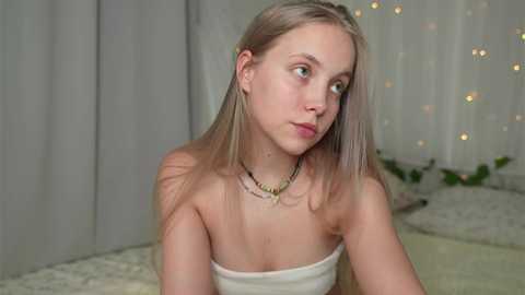 Media: Video of a young Caucasian woman with straight, platinum blonde hair, wearing a strapless, off-white dress, and a gold chain necklace, looking contemplative in a softly lit, cozy bedroom with fairy lights and white curtains.