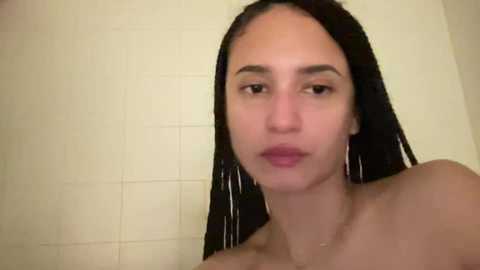 Media: Video of a young, light-skinned woman with long, straight black hair, wearing large silver hoop earrings, taking a shower in a tiled bathroom. She has a neutral expression and is topless, with water droplets on her skin.