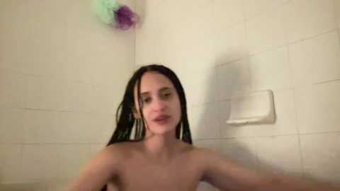 Media: A video of a nude young woman with wet, shoulder-length black hair, standing in a white-tiled shower, holding a colorful shower puff.