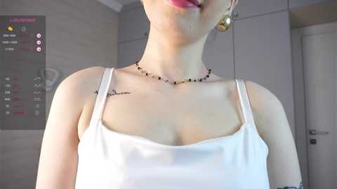 Media: A video of a fair-skinned woman with light pink lipstick, wearing a white spaghetti-strap tank top, gold earrings, and a necklace, in a modern, light-toned room.