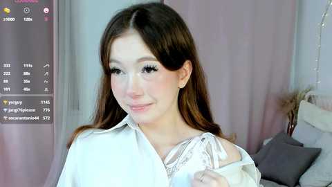 Media: A video of a young, fair-skinned woman with long brown hair, wearing a white shirt, smiling softly. The background shows a bed with a grey pillow, and a pink curtain.