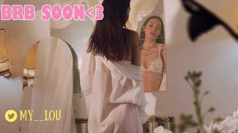 Media: Video: A woman with long dark hair, in a white robe, looks at herself in a mirror. Soft, warm lighting. Text: \"BBB SOKE\" and \"MY/LOU\" in the top-left and bottom-left, respectively.