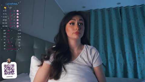 Media: A video of a young woman with long dark hair, wearing a white t-shirt, sitting in a dimly lit bedroom with blue curtains.