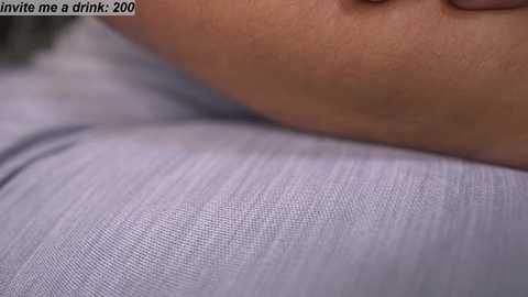 Media: Video of a close-up of a person's upper arm and hand, showcasing a light purple shirt with fine fabric texture. The background is blurred, with the text \"invite me a drink. 200\" visible in the top left corner.