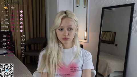 A video of a blonde woman with long hair, wearing a white T-shirt with a pink heart, sitting in a modern bedroom with a mirror, bed, and window with beige curtains.