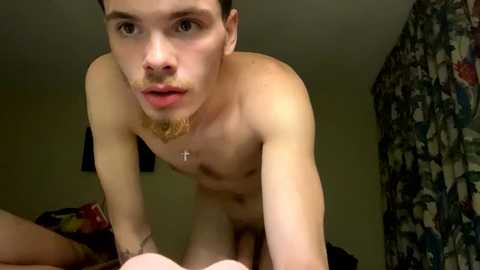 Media: A video of a young, fair-skinned, clean-shaven man with short brown hair, wearing only a necklace, kneeling naked in a bedroom with patterned curtains and a bed in the background.