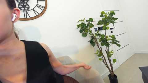 Media: Video of a woman with fair skin, wearing a black sleeveless top, seated at a table, listening to white earbuds, with a small green plant in the background.