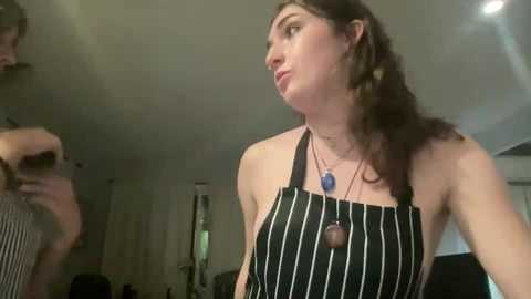 Media: A candid video of a woman with curly brown hair wearing a black-and-white striped apron, standing indoors in a dimly lit room.