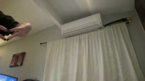 Media: Video of a person in a white T-shirt leaning over a bed with white curtains, air conditioner, and a framed picture on a beige wall.