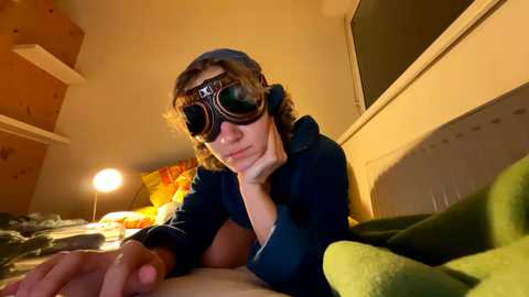 Media: Video of a young person with long hair, wearing dark glasses and a blue hoodie, lying on a bed in a dimly lit room with wooden shelves and a yellow lamp.