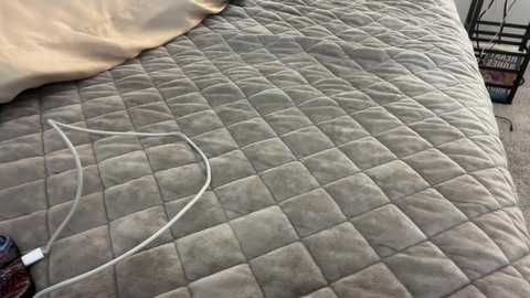 Media: Video of a beige, quilted bedspread with a white charging cable snaking across it, and a beige pillow visible at the top left corner. The background shows a blurred, modern bedroom with a black metal frame.