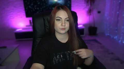 Media: Video of a young woman with long, straight red hair, wearing a black sequined shirt, sitting in a black leather chair in a dimly lit room with purple and blue LED lights.