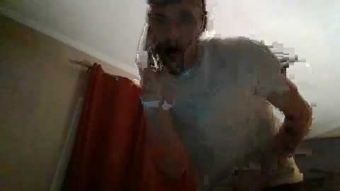 Media: Video of a man with a medium build, wearing a white T-shirt, leaning against a wall with a red curtain in a dimly lit room. His facial expression is obscured due to a glitchy image.