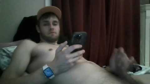 Media: Video of a shirtless, fair-skinned, young man with a light beard, wearing a baseball cap, looking at a smartphone. He has a wristwatch and lies on a bed with a red curtain background.