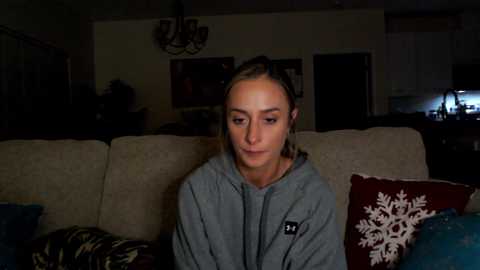 Media: Video of a young woman with light skin, brown hair, and a slim build, sitting on a beige couch in a dimly lit living room. She wears a grey hoodie.